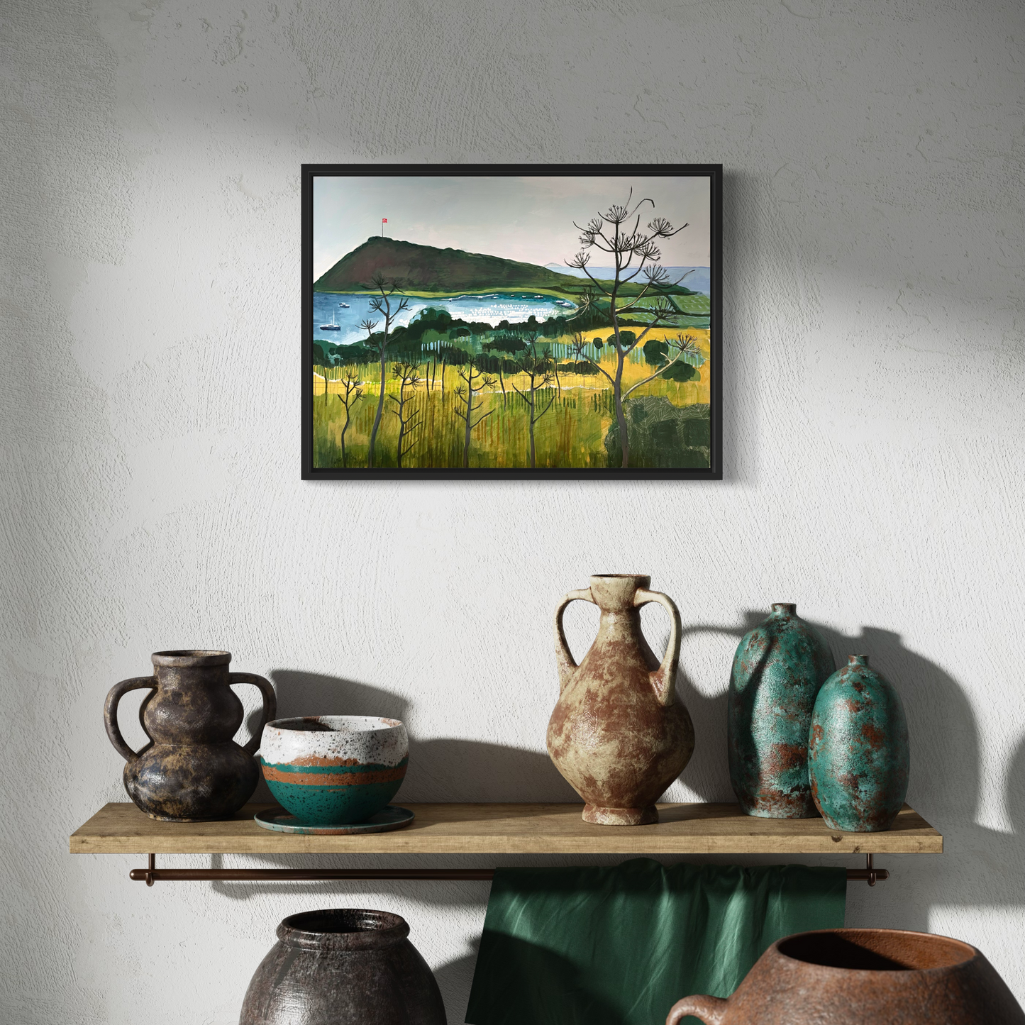 Aegean Autumn landscape by Elaine Boylan. View of Gümüşlük bay on the Bodrum Peninsula in Turkey. Pale blue sky, turquoise sea, hill with flag pole flying the Turkish flag, field in the foreground. Colours are mainly greens, golds and blues. High quality giclée print on stretched canvas with black float frame. Print is shown in a room above a shelf with pottery.