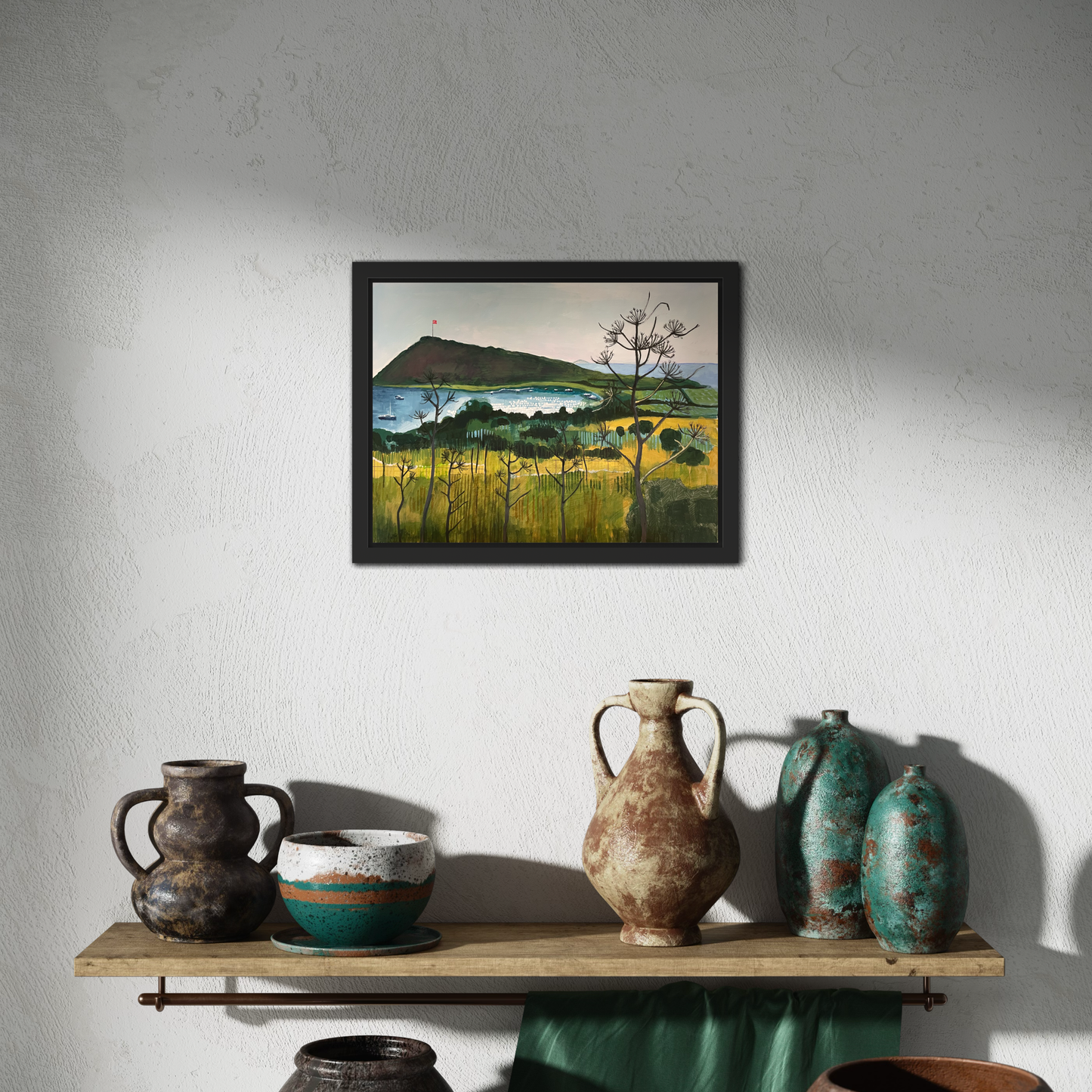 Aegean Autumn mixed media original landscape by Elaine Boylan. Black wood tray frame. View of Gümüşlük bay on the Bodrum Peninsula in Turkey. Pale blue sky, turquoise sea, hill with flag pole flying the Turkish flag, field in the foreground. Colours are mainly greens, golds and blues. Materials are collage and acrylic paint on board. Painting is shown in a room above a shelf with a selection of pottery.