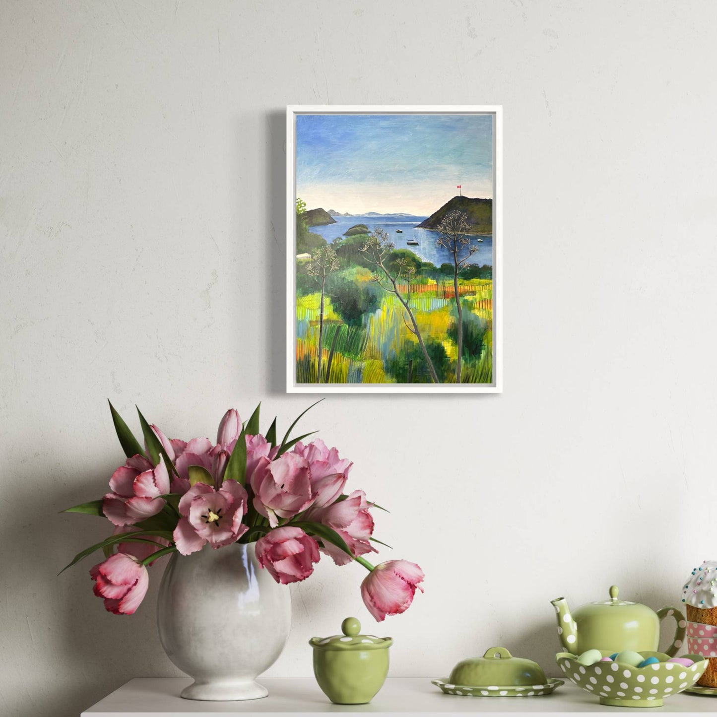 After the Rain landscape by Elaine Boylan. View across Gümüşlük bay on the Bodrum Peninsula in Turkey. Pale blue sky, turquoise sea, hill with flag pole flying the Turkish flag, field in the foreground. Colours are mainly greens, golds and blues. High quality giclée print on stretched canvas framed in a white float frame. Print is shown in a bright room above a console table with a vase of tulips, and pale green crockery.