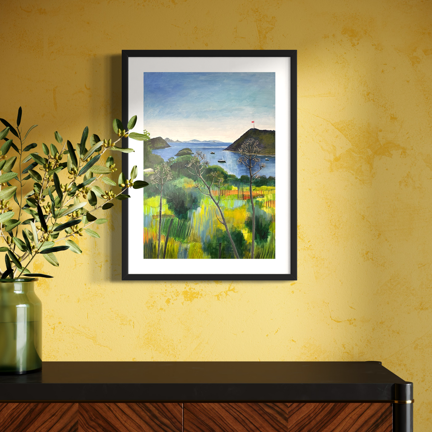 After the Rain landscape by Elaine Boylan. View from Gümüşlük on the Bodrum Peninsula in Turkey. Pale blue sky over the Aegean sea, hill with flag pole flying the Turkish flag, field in the foreground, Greek islands in the distance. Colours are mainly greens, golds and blues. High quality giclée print on fine art paper with mount/mat and black frame. Print is shown in a bright room on a yellow wall above a sideboard with a vase.