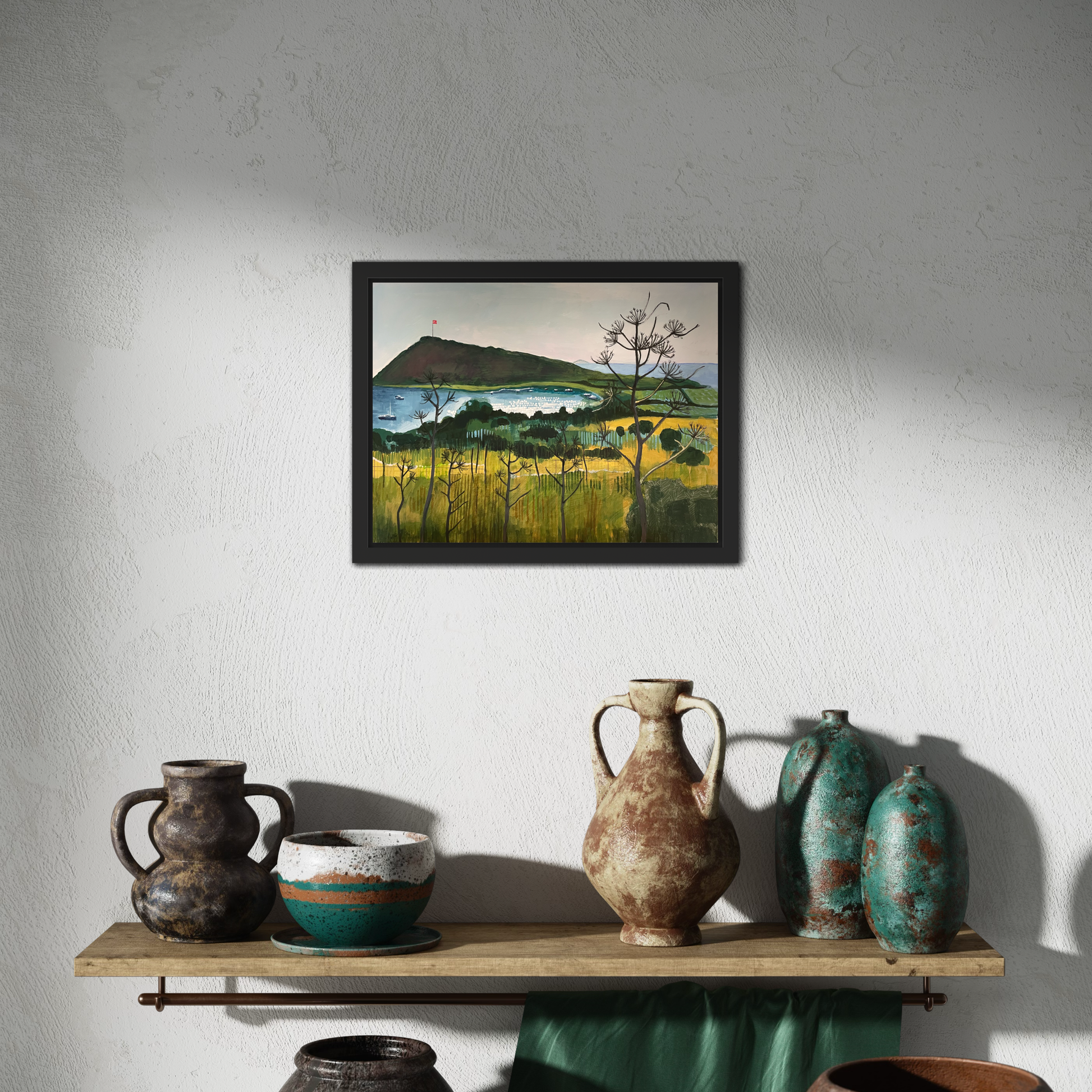 Aegean Autumn mixed media original landscape by Elaine Boylan. Black wood tray frame. View of Gümüşlük bay on the Bodrum Peninsula in Turkey. Pale blue sky, turquoise sea, hill with flag pole flying the Turkish flag, field in the foreground. Colours are mainly greens, golds and blues. Materials are collage and acrylic paint on board. Painting is shown in a room above a shelf with a selection of pottery. 