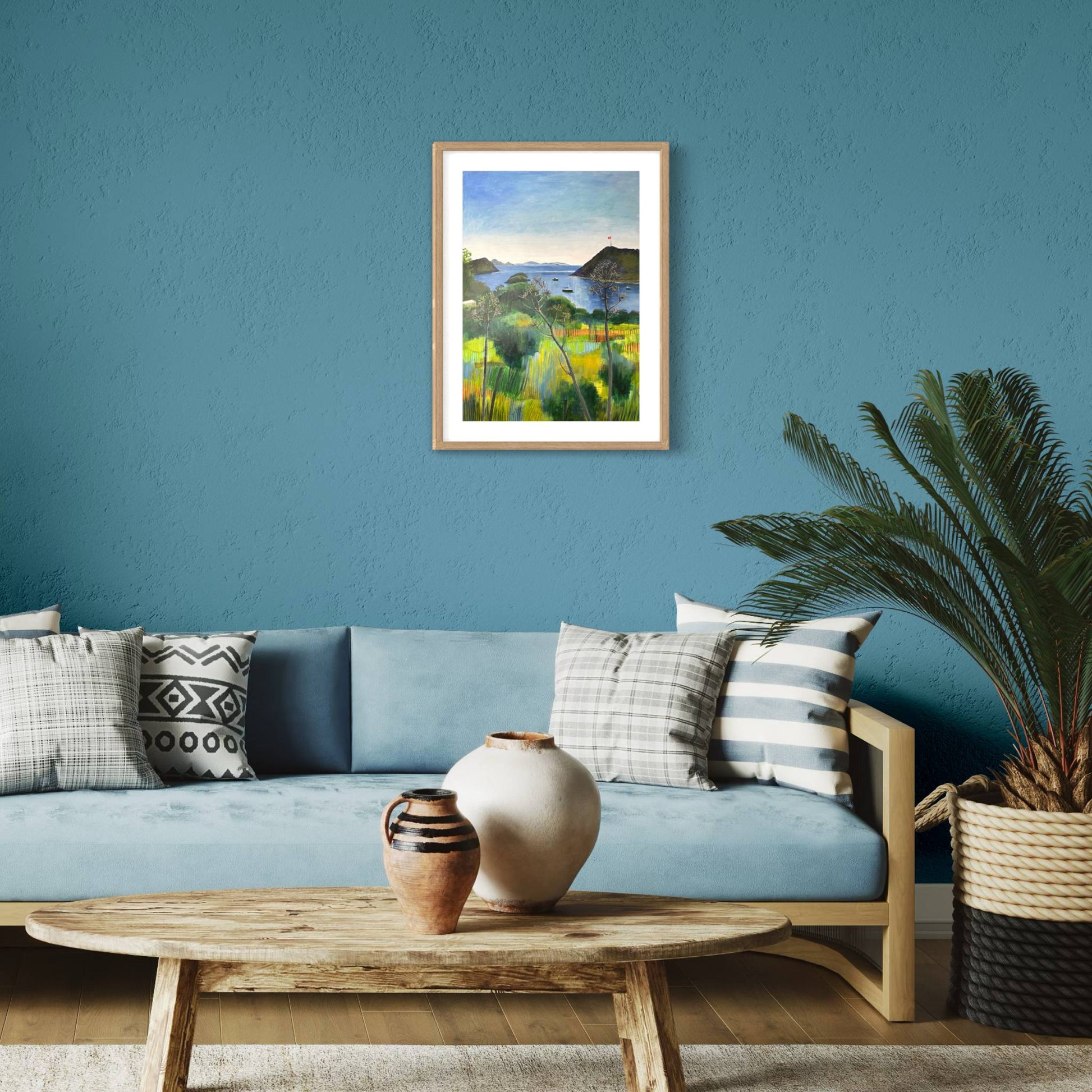 After the Rain landscape by Elaine Boylan. View from Gümüşlük on the Bodrum Peninsula in Turkey. Pale blue sky over the Aegean sea, hill with flag pole flying the Turkish flag, field in the foreground, Greek islands in the distance. Colours are mainly greens, golds and blues. High quality giclée print on fine art paper with mount/mat and natural wood frame. Print is shown in a contemporary style room on a teal wall above a pale blue sofa.